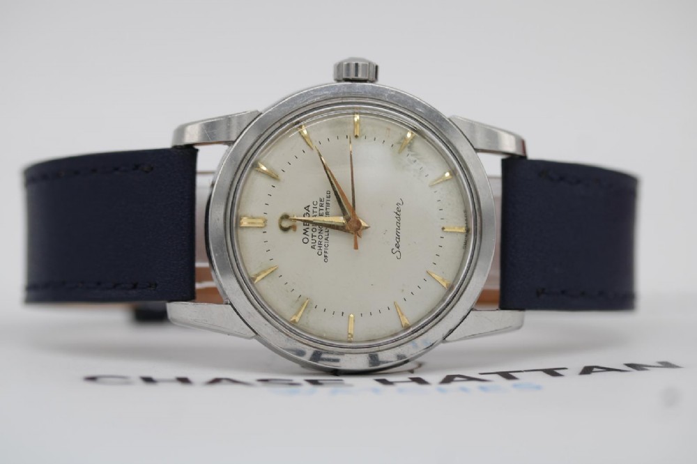 A vintage 1956 Omega Seamaster automatic wristwatch featuring a silver-coloured stainless steel case, off-white dial with gold-tone markers, and a navy blue leather strap. View IX