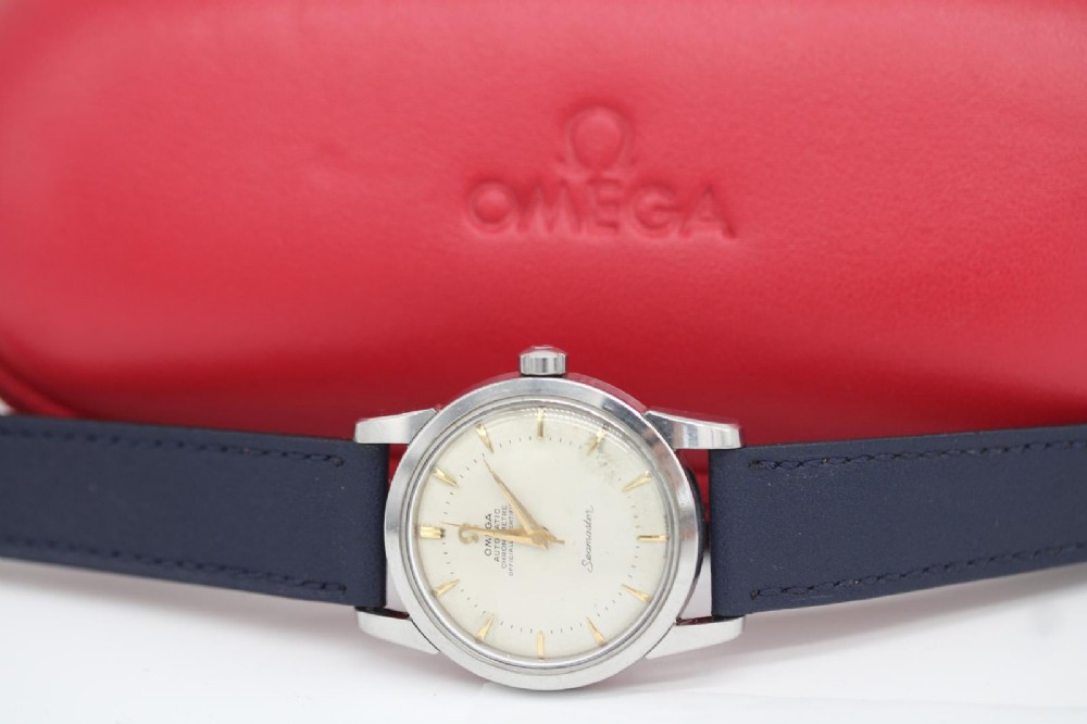 A vintage 1956 Omega Seamaster automatic wristwatch featuring a silver-coloured stainless steel case, off-white dial with gold-tone markers, and a navy blue leather strap. View XI