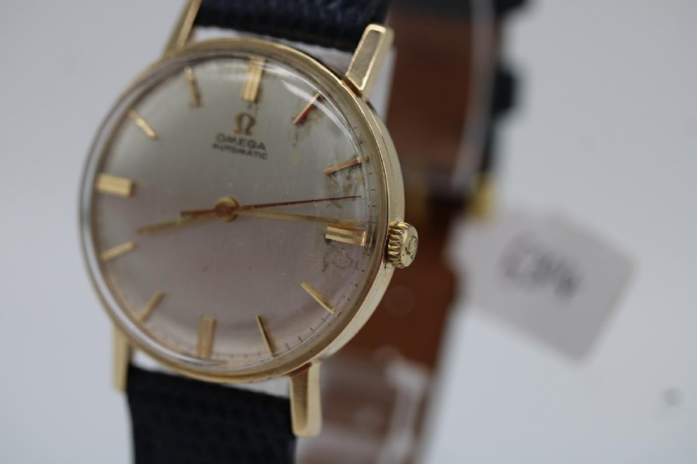 A 1962 Omega automatic wristwatch featuring a round 9ct gold case, silver dial with baton indices, and a blue textured leather strap. View VI