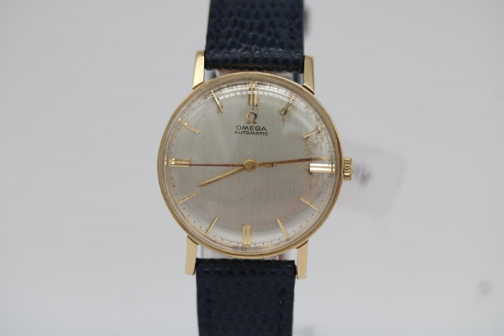 A 1962 Omega automatic wristwatch featuring a round 9ct gold case, silver dial with baton indices, and a blue textured leather strap.