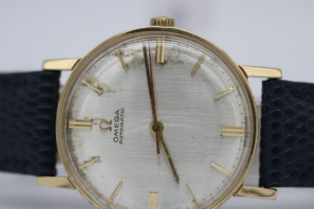 A 1962 Omega automatic wristwatch featuring a round 9ct gold case, silver dial with baton indices, and a blue textured leather strap. View IX