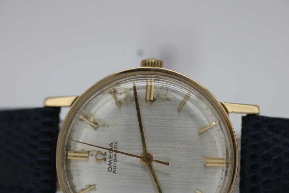 A 1962 Omega automatic wristwatch featuring a round 9ct gold case, silver dial with baton indices, and a blue textured leather strap. View X