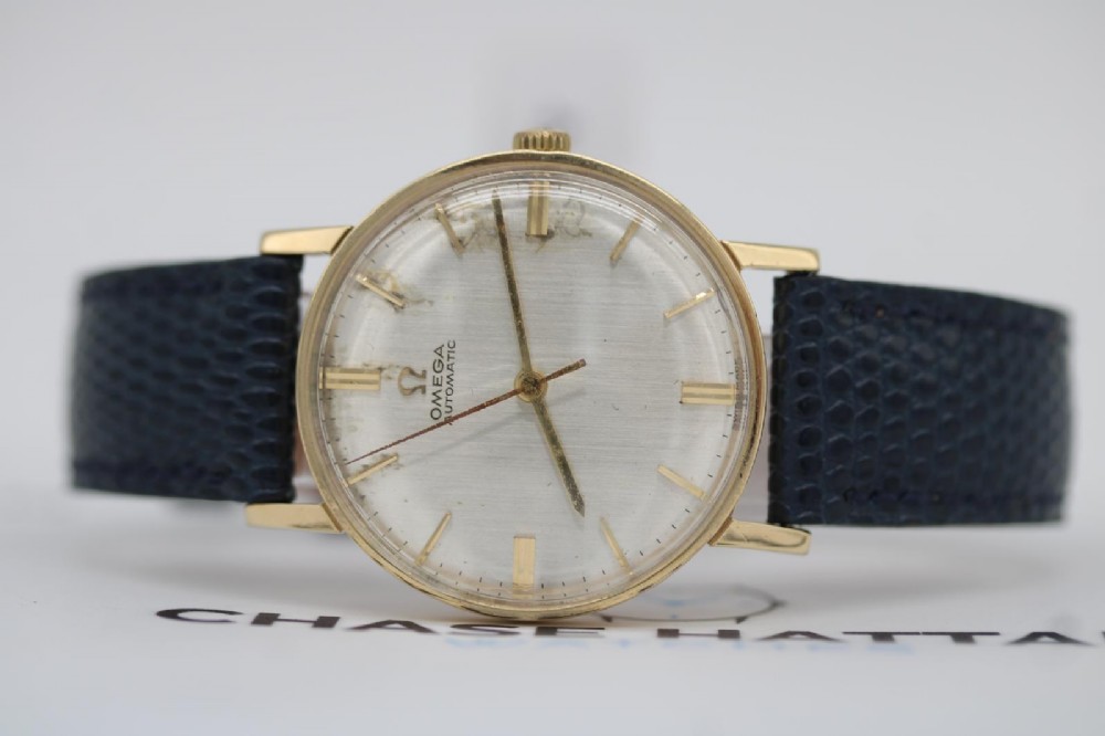 A 1962 Omega automatic wristwatch featuring a round 9ct gold case, silver dial with baton indices, and a blue textured leather strap. View XI