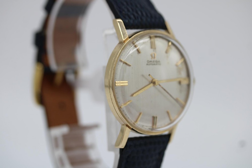 A 1962 Omega automatic wristwatch featuring a round 9ct gold case, silver dial with baton indices, and a blue textured leather strap. View XIII