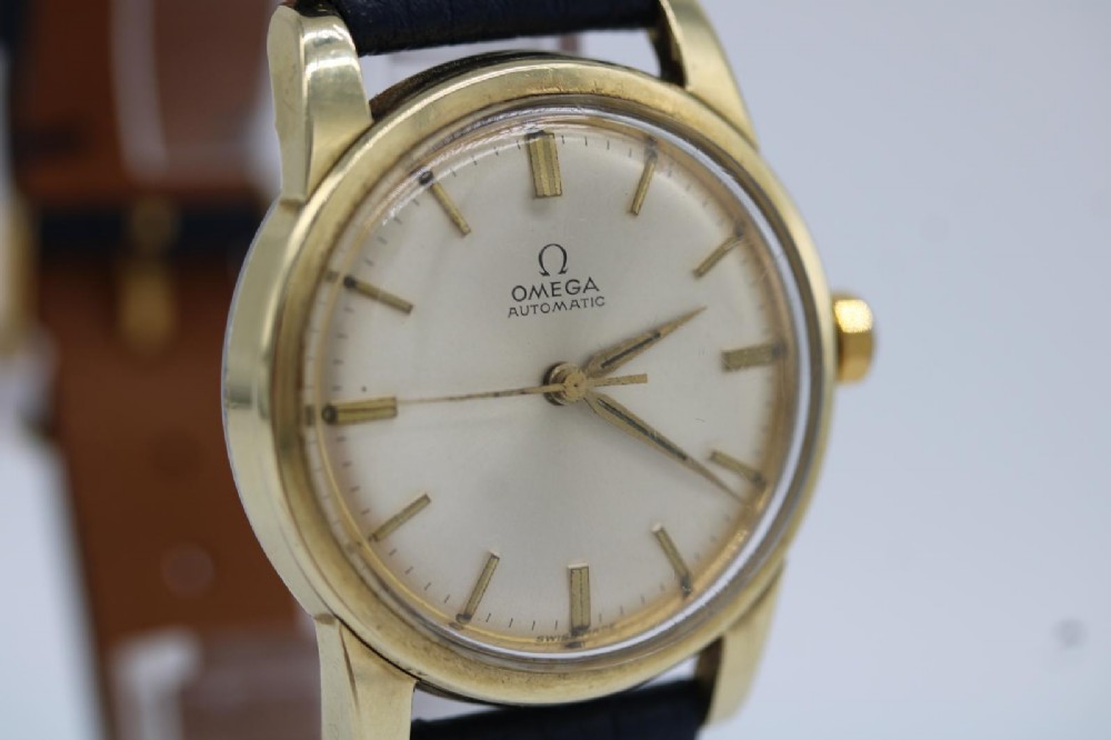 A gold-plated circular vintage wristwatch featuring a cream dial, gold hour indices, and a dark blue leather strap. View VII