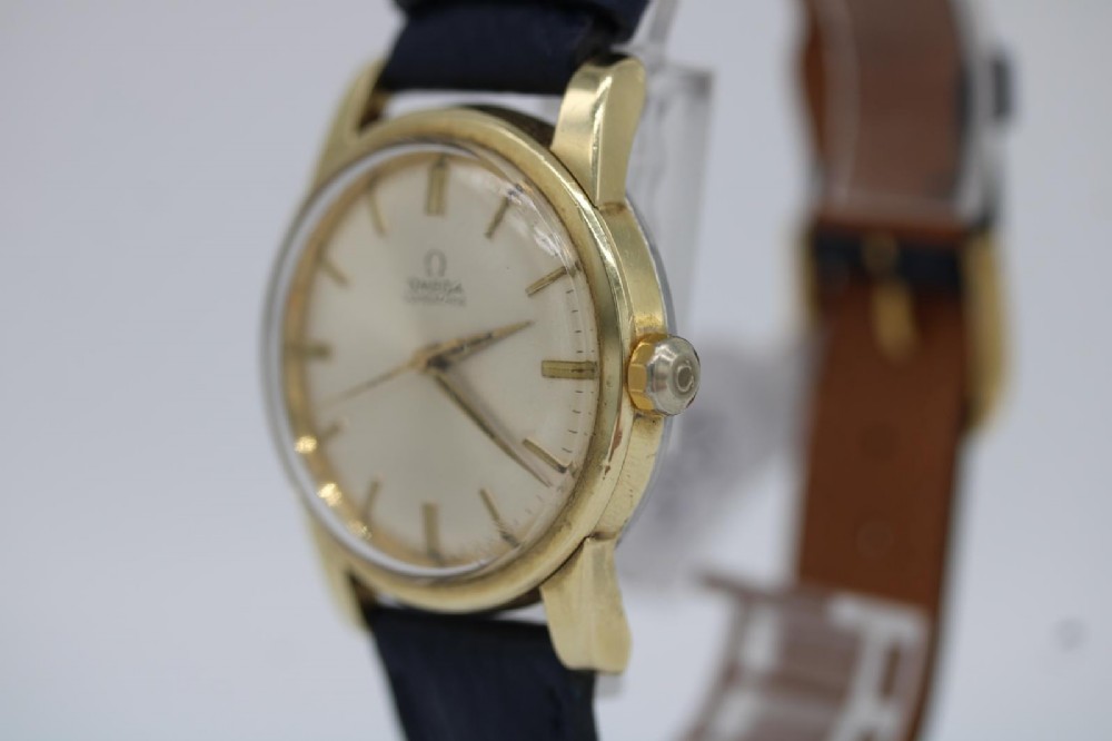 A gold-plated circular vintage wristwatch featuring a cream dial, gold hour indices, and a dark blue leather strap. View X
