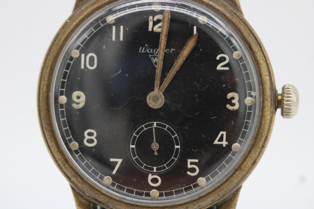 A close-up photograph of a 1940s Wagner German military wristwatch featuring a black dial, luminous Arabic numerals, a small sub-seconds dial, and a patinated brass case.