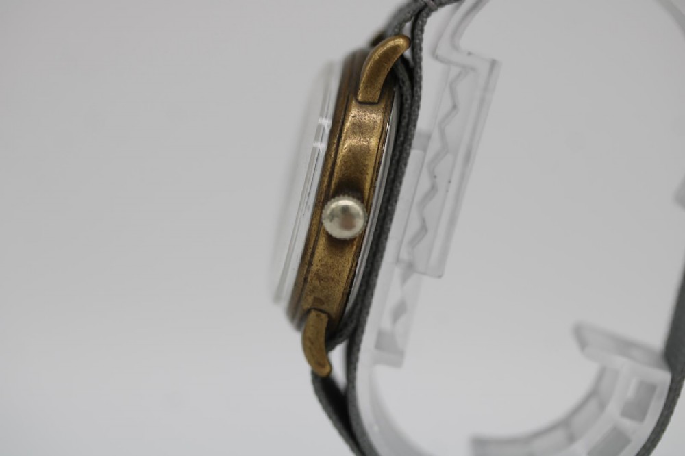 A close-up photograph of a 1940s Wagner German military wristwatch featuring a black dial, luminous Arabic numerals, a small sub-seconds dial, and a patinated brass case. View III