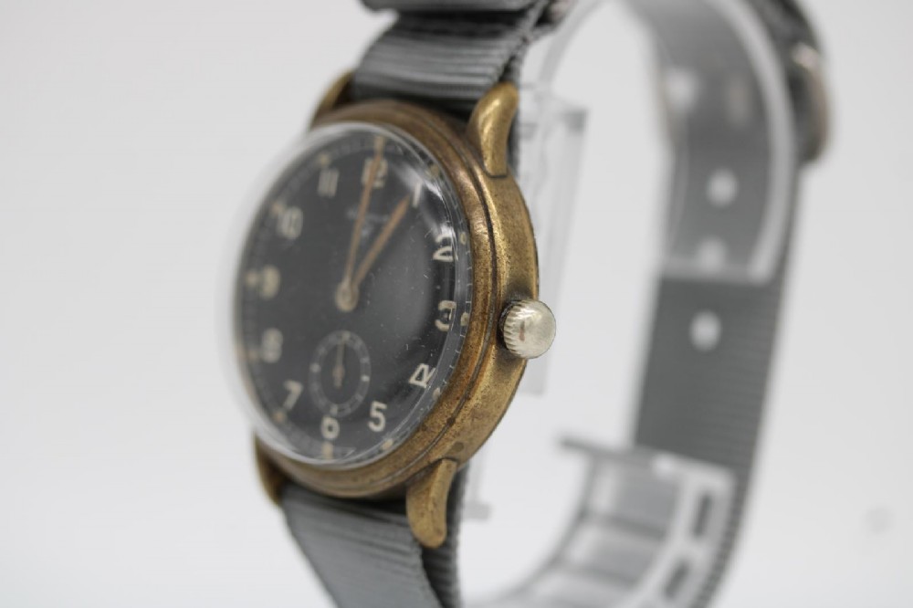 A close-up photograph of a 1940s Wagner German military wristwatch featuring a black dial, luminous Arabic numerals, a small sub-seconds dial, and a patinated brass case. View IV