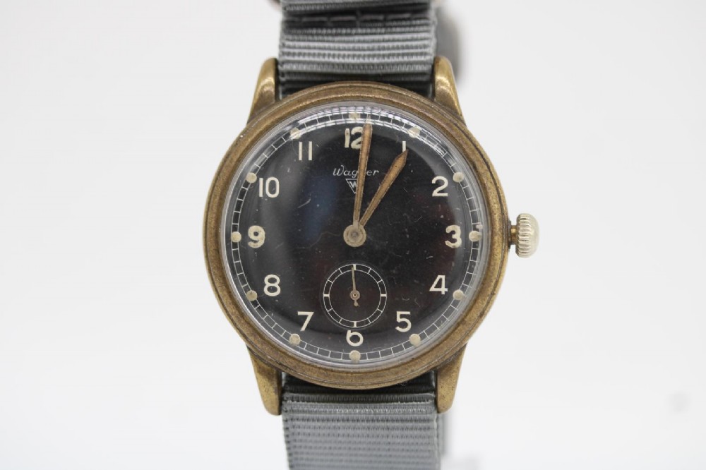 A close-up photograph of a 1940s Wagner German military wristwatch featuring a black dial, luminous Arabic numerals, a small sub-seconds dial, and a patinated brass case. View V