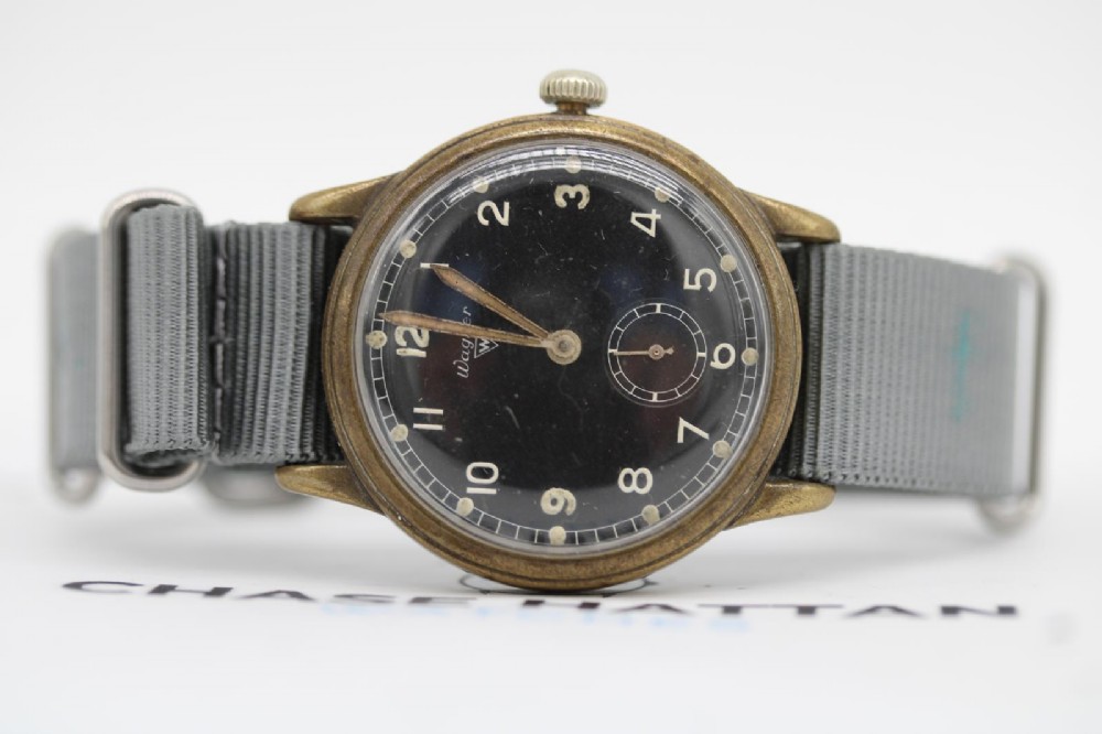 A close-up photograph of a 1940s Wagner German military wristwatch featuring a black dial, luminous Arabic numerals, a small sub-seconds dial, and a patinated brass case. View VI