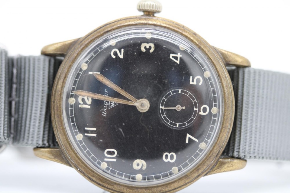 A close-up photograph of a 1940s Wagner German military wristwatch featuring a black dial, luminous Arabic numerals, a small sub-seconds dial, and a patinated brass case. View VIII
