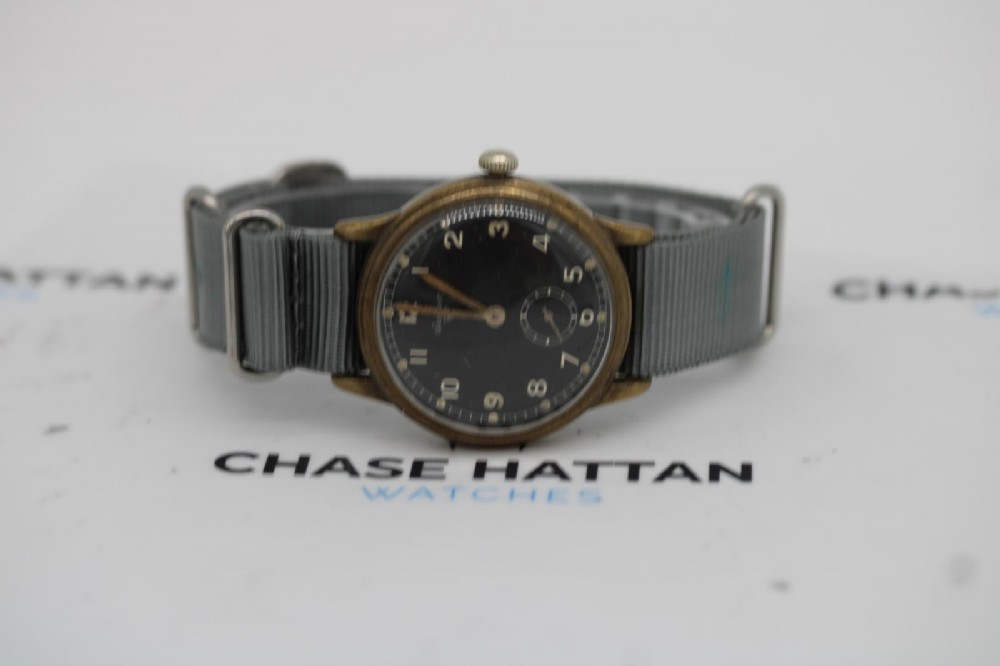 A close-up photograph of a 1940s Wagner German military wristwatch featuring a black dial, luminous Arabic numerals, a small sub-seconds dial, and a patinated brass case. View IX