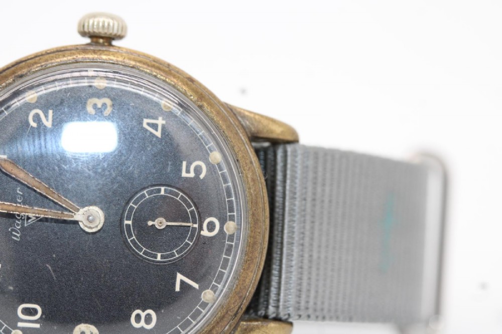 A close-up photograph of a 1940s Wagner German military wristwatch featuring a black dial, luminous Arabic numerals, a small sub-seconds dial, and a patinated brass case. View X