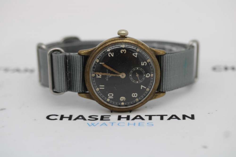 A close-up photograph of a 1940s Wagner German military wristwatch featuring a black dial, luminous Arabic numerals, a small sub-seconds dial, and a patinated brass case. View XI
