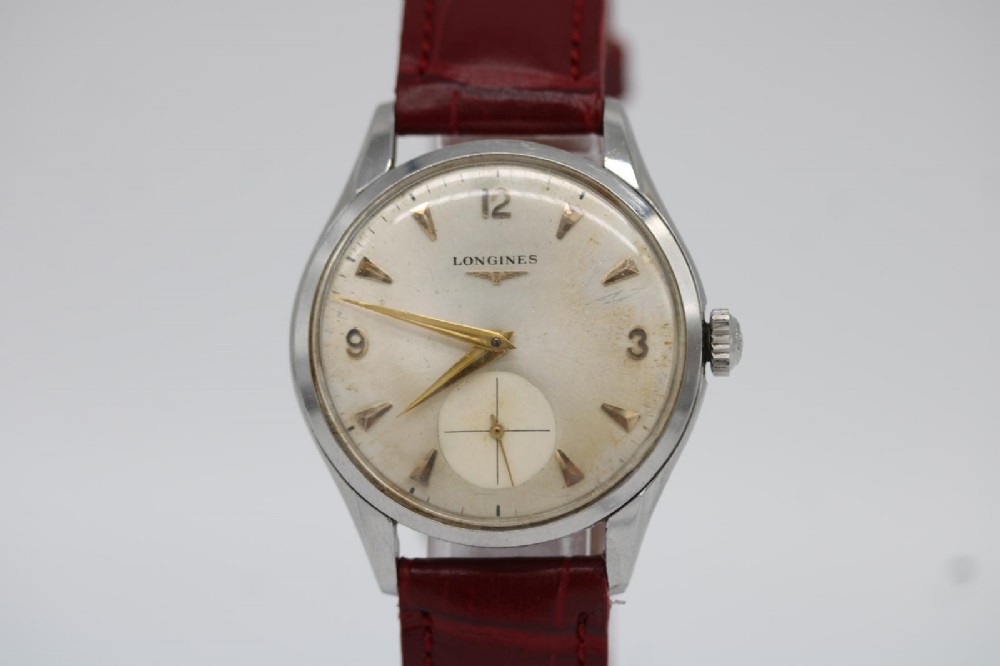 A 1950s Longines wristwatch with a stainless steel case, off-white dial with gold-tone hour markers, sub-seconds dial, and a burgundy leather strap.