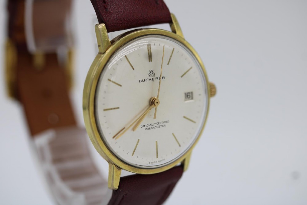 A circular vintage Bucherer gold-plated automatic wristwatch featuring a silver dial, gold hour indices, a date aperture at three oclock, and a burgundy leather strap. View II