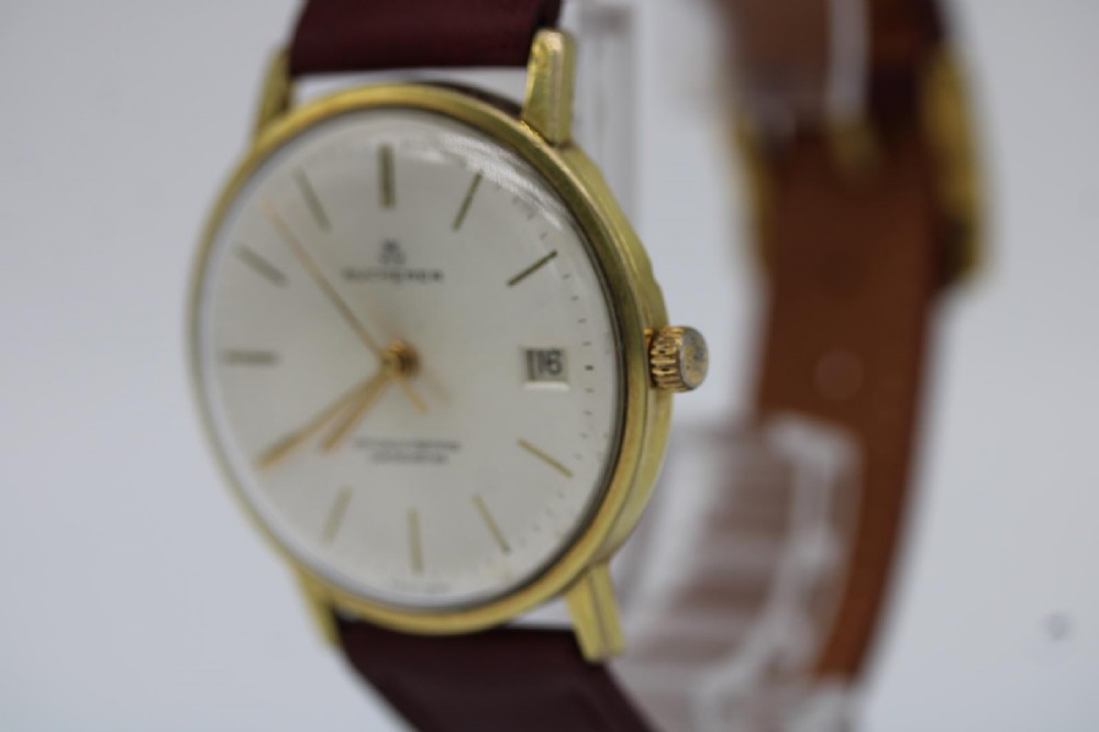 A circular vintage Bucherer gold-plated automatic wristwatch featuring a silver dial, gold hour indices, a date aperture at three oclock, and a burgundy leather strap. View III