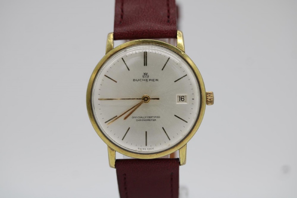 A circular vintage Bucherer gold-plated automatic wristwatch featuring a silver dial, gold hour indices, a date aperture at three oclock, and a burgundy leather strap.