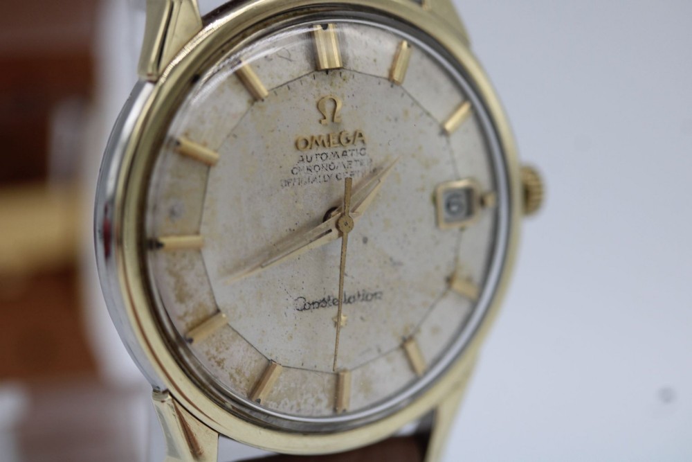 A vintage gold-plated Omega Constellation wristwatch featuring a faceted pie-pan dial, date window, and a brown leather strap. View V