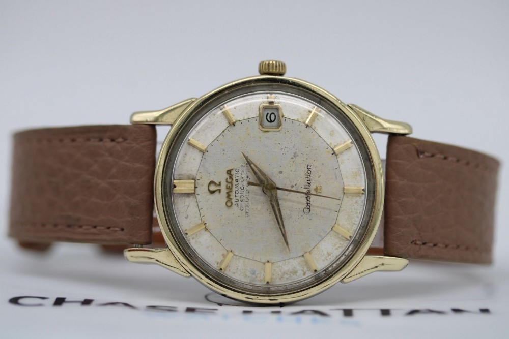 A vintage gold-plated Omega Constellation wristwatch featuring a faceted pie-pan dial, date window, and a brown leather strap. View X