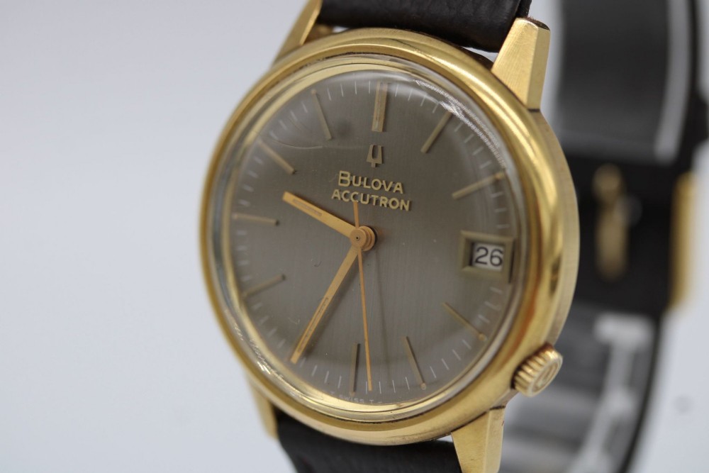 A 1960s circular gold-plated Bulova Accutron wristwatch with a grey-brown dial, date display, and brown leather strap. View II