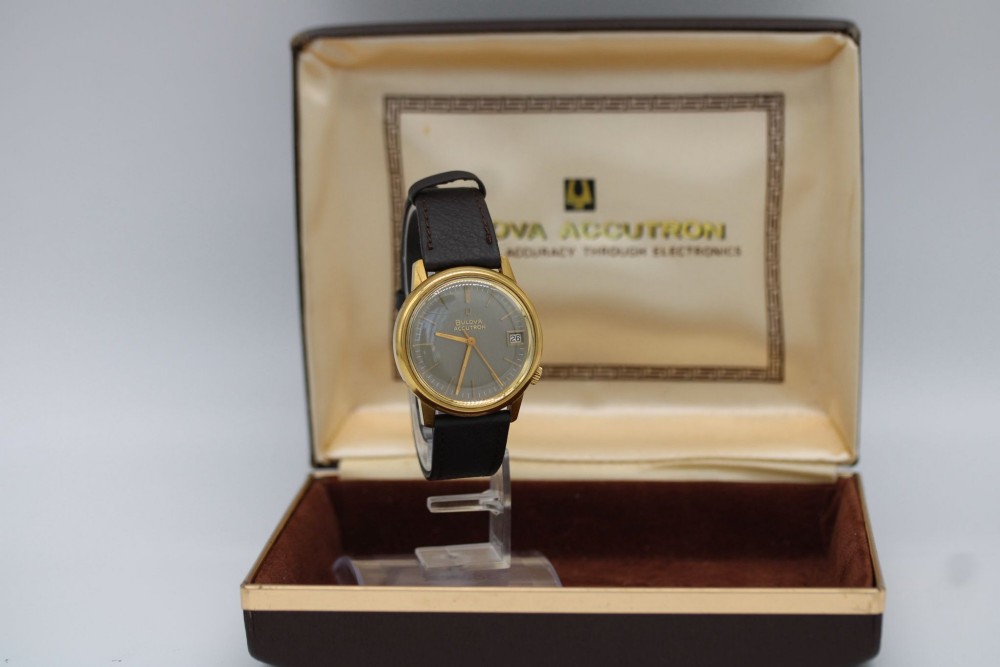 A 1960s circular gold-plated Bulova Accutron wristwatch with a grey-brown dial, date display, and brown leather strap. View III