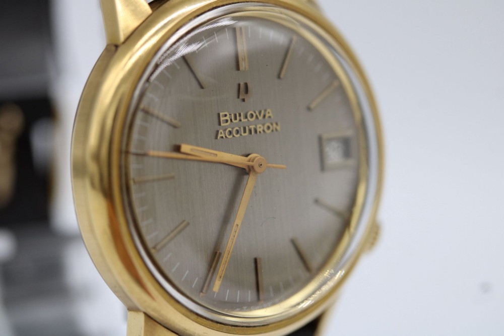 A 1960s circular gold-plated Bulova Accutron wristwatch with a grey-brown dial, date display, and brown leather strap. View IV