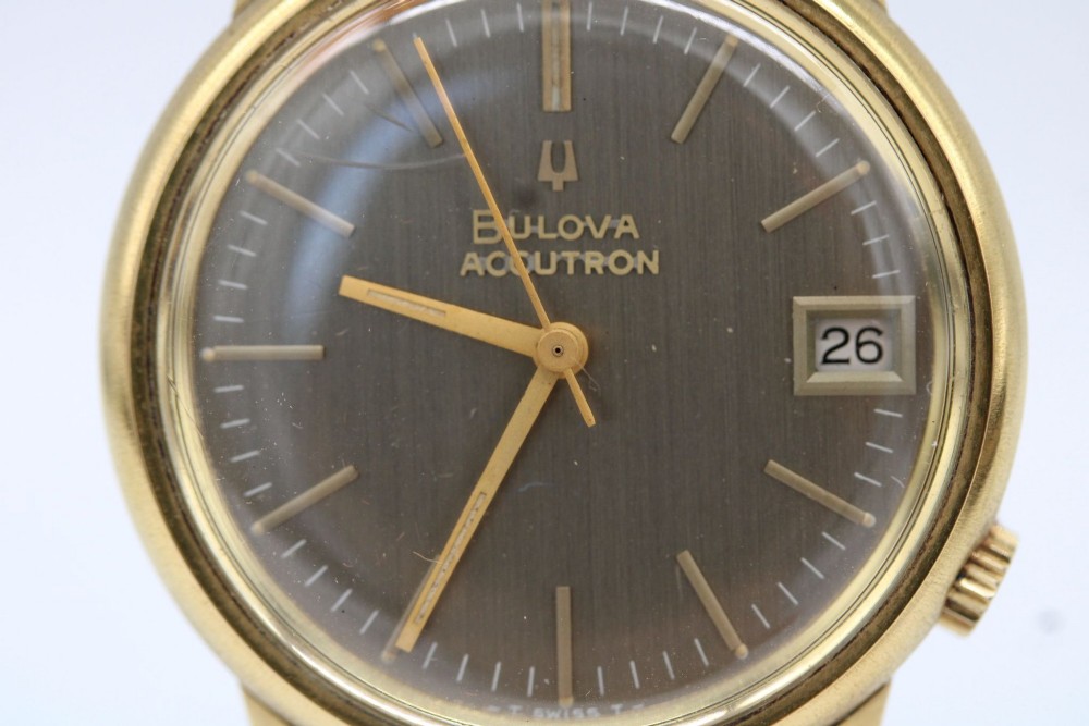 A 1960s circular gold-plated Bulova Accutron wristwatch with a grey-brown dial, date display, and brown leather strap. View V
