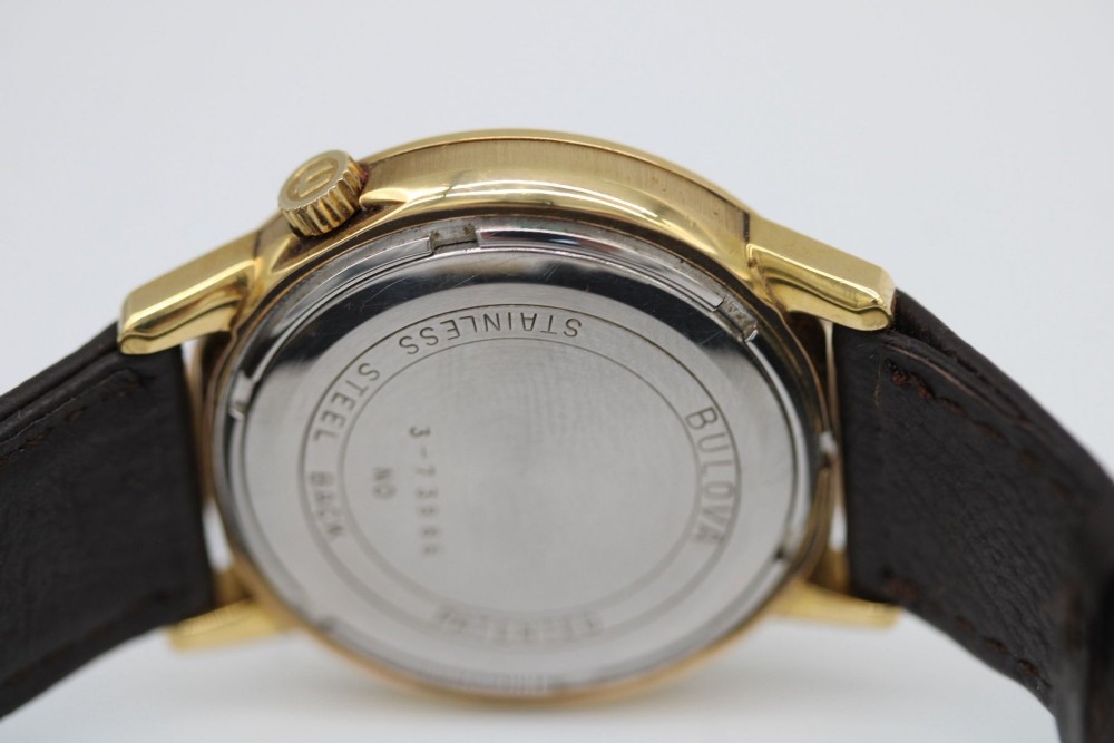 A 1960s circular gold-plated Bulova Accutron wristwatch with a grey-brown dial, date display, and brown leather strap. View VII