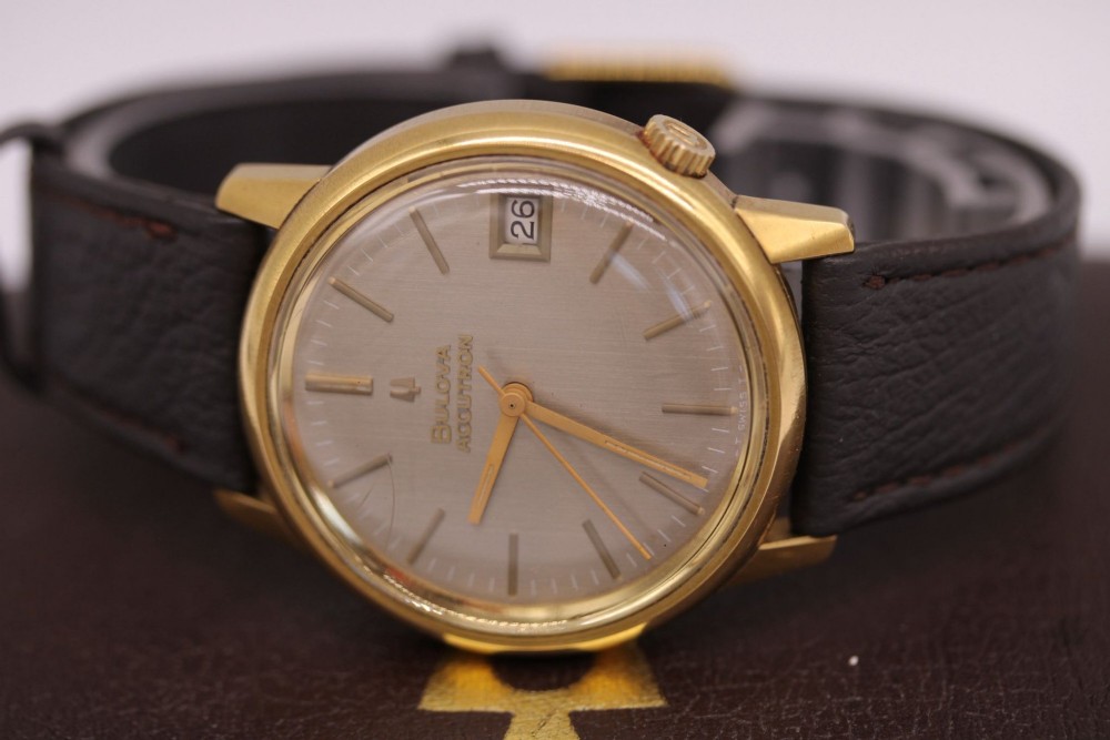 A 1960s circular gold-plated Bulova Accutron wristwatch with a grey-brown dial, date display, and brown leather strap. View IX