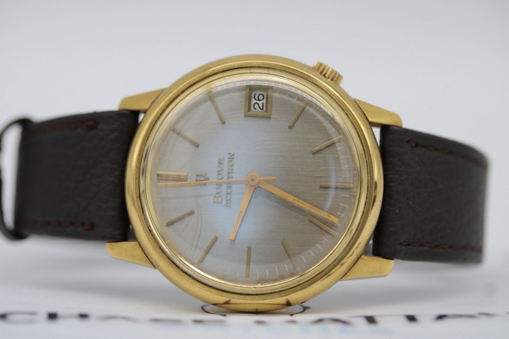 A 1960s circular gold-plated Bulova Accutron wristwatch with a grey-brown dial, date display, and brown leather strap. View X