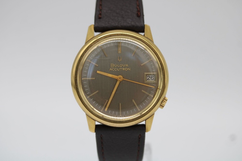 A 1960s circular gold-plated Bulova Accutron wristwatch with a grey-brown dial, date display, and brown leather strap.