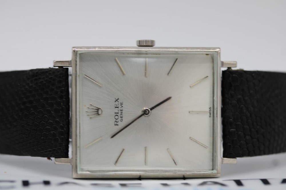 A 1960s square Rolex Cellini wristwatch featuring an 18ct white gold case, silver sunburst dial with simple index markers, and a black textured leather strap. View XI