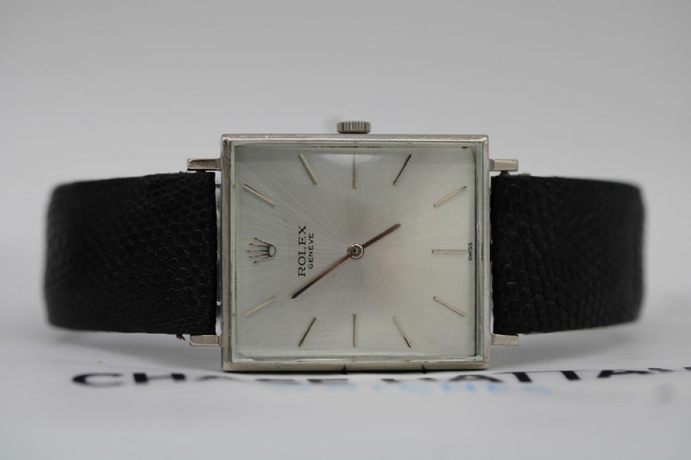 A 1960s square Rolex Cellini wristwatch featuring an 18ct white gold case, silver sunburst dial with simple index markers, and a black textured leather strap. View XII