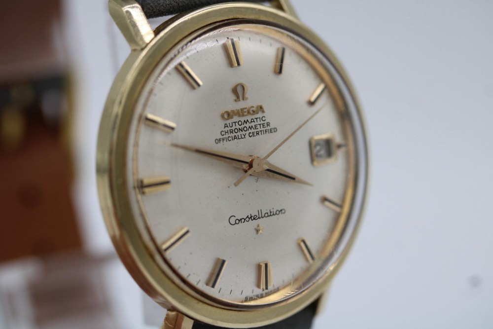A vintage Omega Constellation wristwatch featuring a gold-plated circular case, silver-tone dial with date aperture, and a grey suede strap. View VII