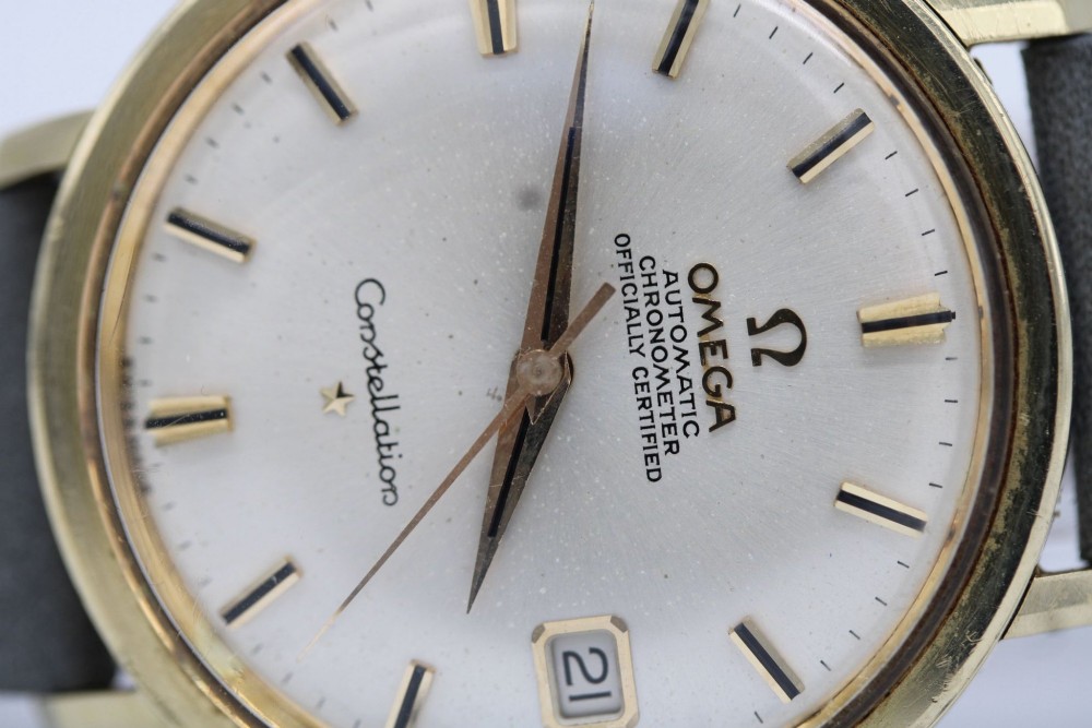 A vintage Omega Constellation wristwatch featuring a gold-plated circular case, silver-tone dial with date aperture, and a grey suede strap. View XI