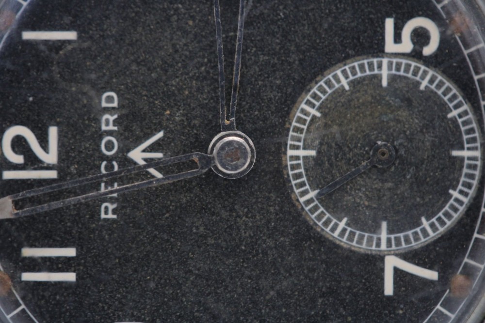 A 1940s Record military wristwatch with a black dial, Arabic numerals, broad arrow mark, and a khaki fabric strap. View V