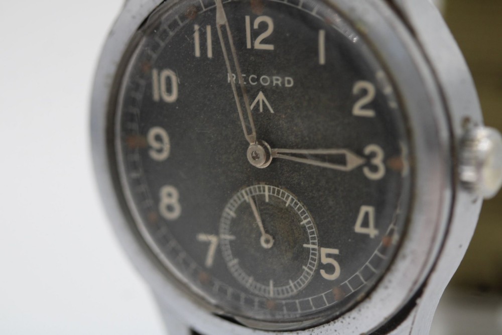A 1940s Record military wristwatch with a black dial, Arabic numerals, broad arrow mark, and a khaki fabric strap. View VII