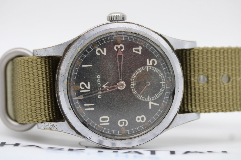 A 1940s Record military wristwatch with a black dial, Arabic numerals, broad arrow mark, and a khaki fabric strap. View XII