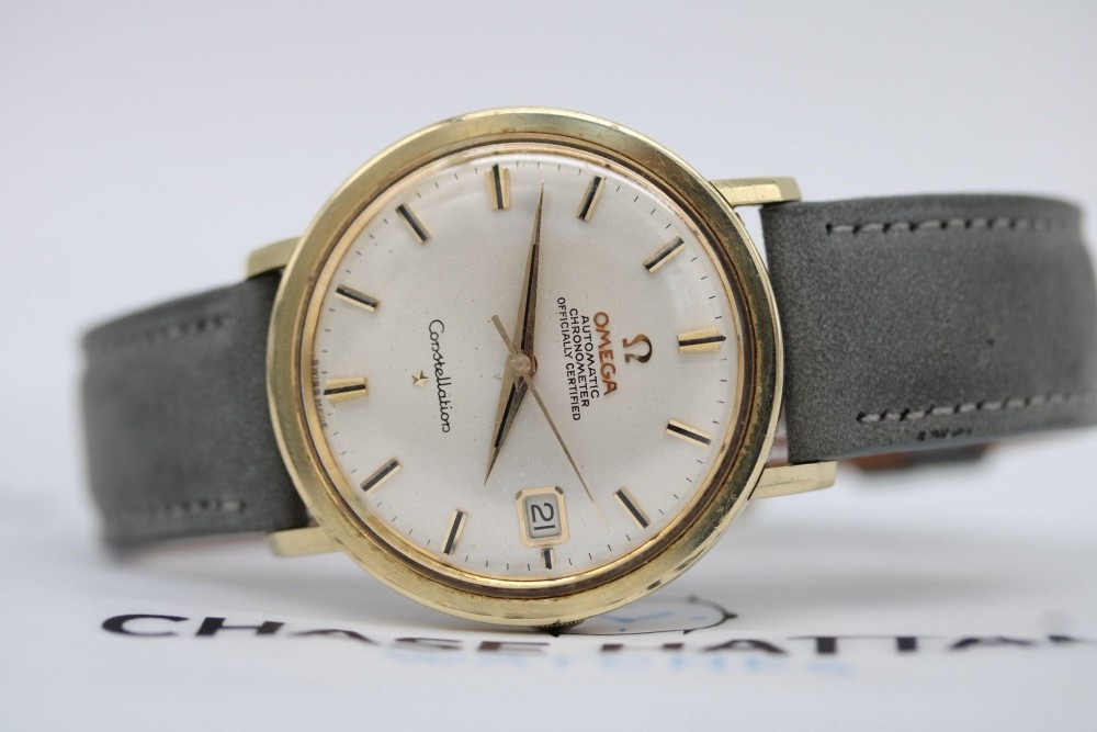 A circular 14ct gold vintage Omega Constellation wristwatch featuring a cream dial with gold baton markers, dauphine hands, date window, and a grey leather strap. View II