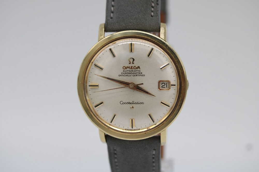 A circular 14ct gold vintage Omega Constellation wristwatch featuring a cream dial with gold baton markers, dauphine hands, date window, and a grey leather strap.