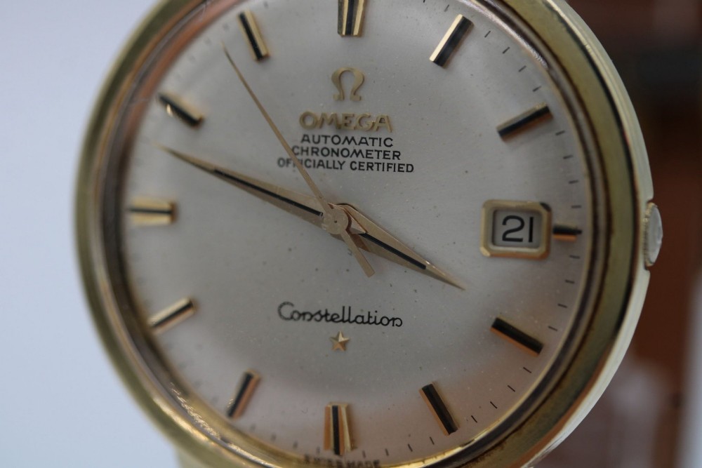 A circular 14ct gold vintage Omega Constellation wristwatch featuring a cream dial with gold baton markers, dauphine hands, date window, and a grey leather strap. View V