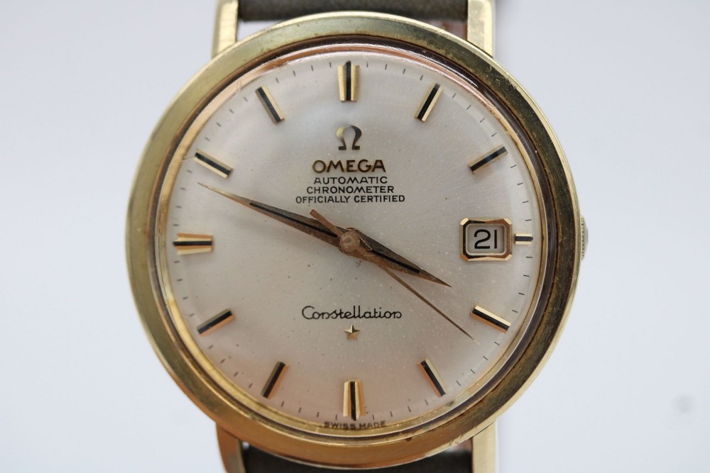 A circular 14ct gold vintage Omega Constellation wristwatch featuring a cream dial with gold baton markers, dauphine hands, date window, and a grey leather strap. View VIII