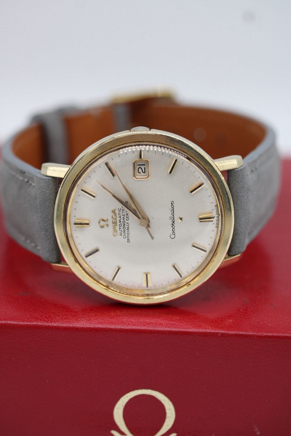 A circular 14ct gold vintage Omega Constellation wristwatch featuring a cream dial with gold baton markers, dauphine hands, date window, and a grey leather strap. View IX