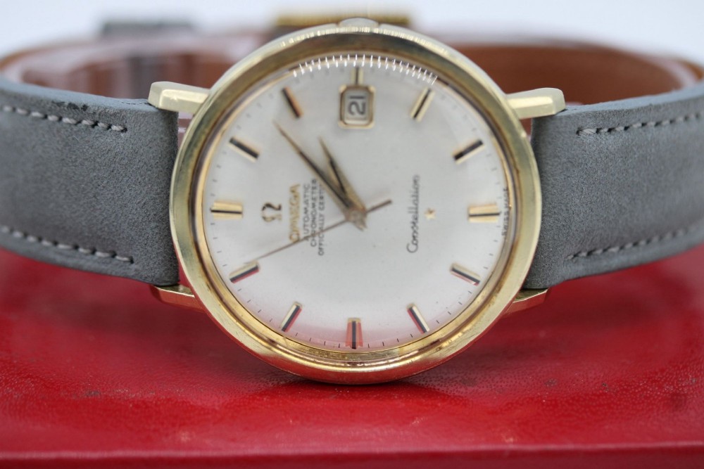 A circular 14ct gold vintage Omega Constellation wristwatch featuring a cream dial with gold baton markers, dauphine hands, date window, and a grey leather strap. View X