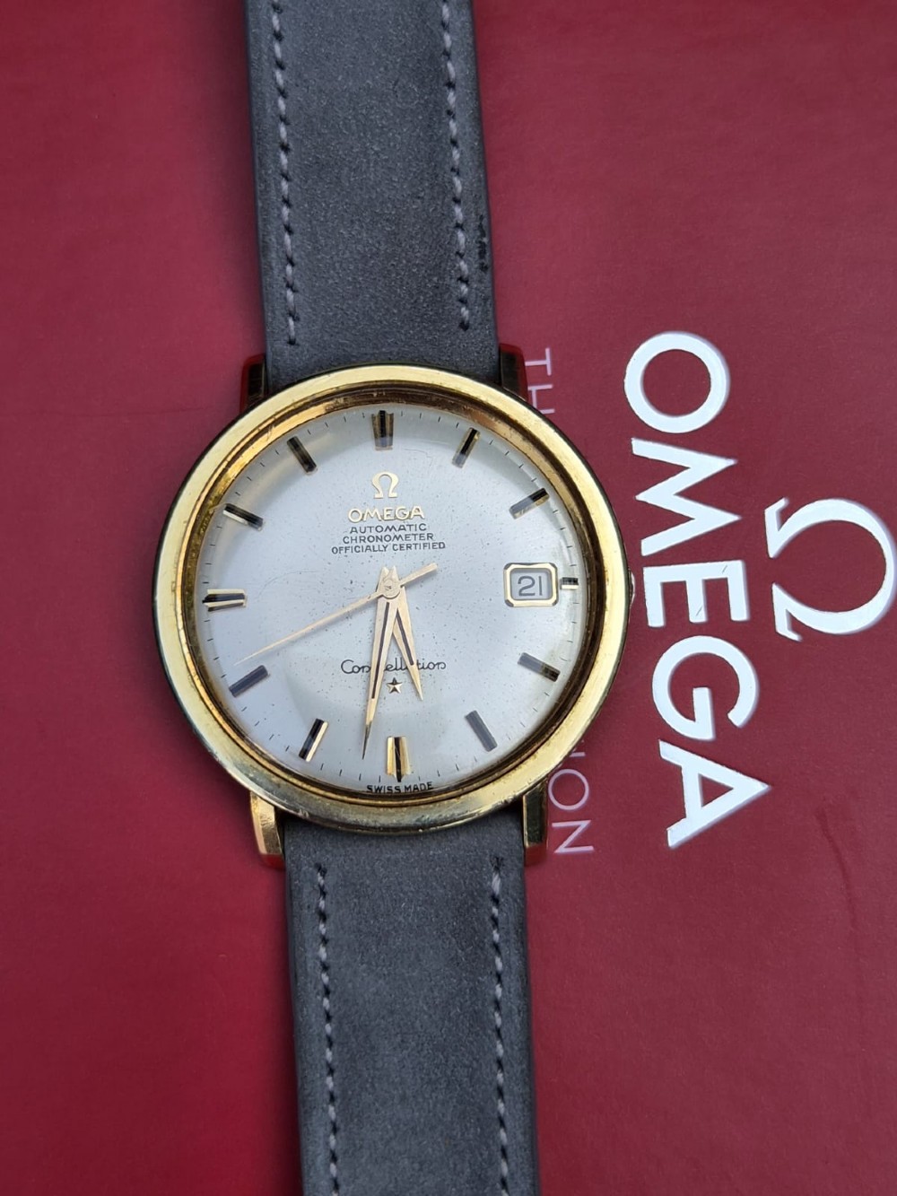 A circular 14ct gold vintage Omega Constellation wristwatch featuring a cream dial with gold baton markers, dauphine hands, date window, and a grey leather strap. View XII