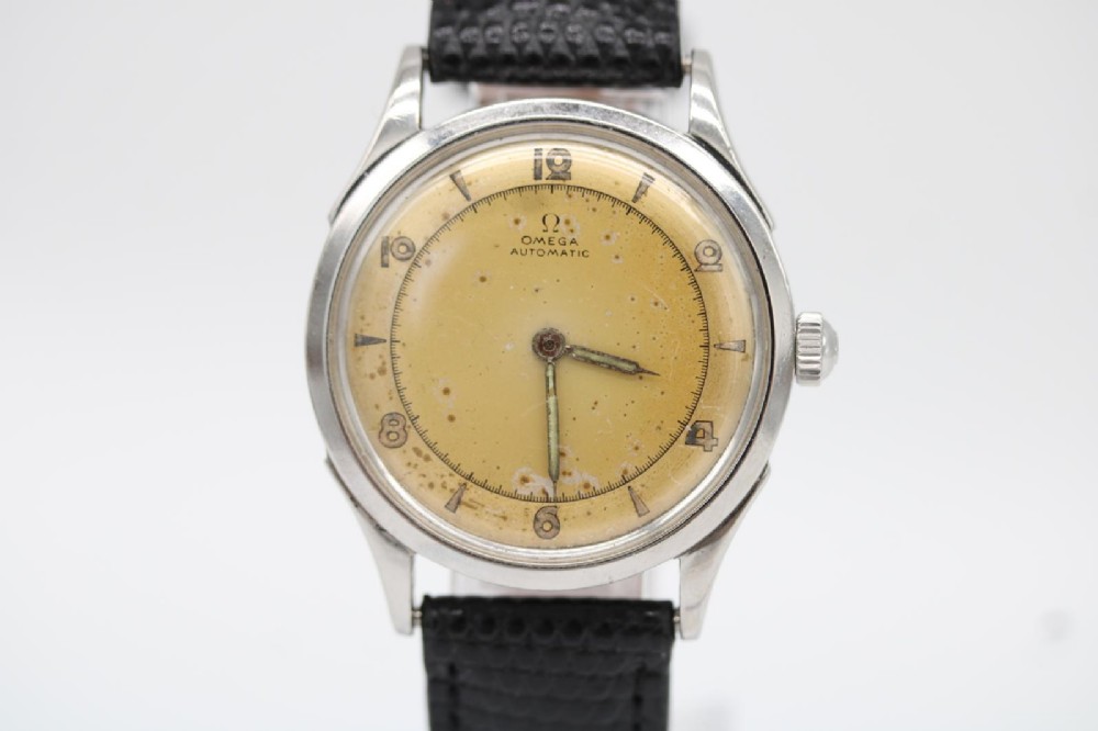 A round 1947 stainless steel Omega wristwatch displaying an aged cream dial with Arabic numerals and fitted with a black textured leather strap.