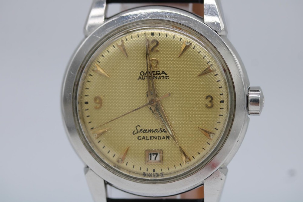 A vintage 1952 Omega Seamaster Calendar wristwatch featuring a stainless steel case, textured gold-coloured dial with date display at six oclock, and a black leather strap. View VI