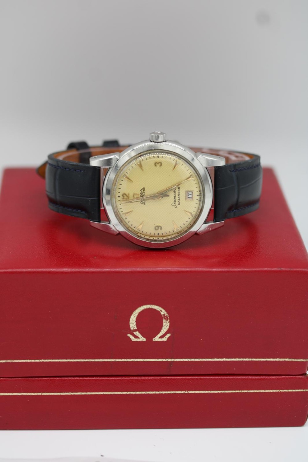 A vintage 1952 Omega Seamaster Calendar wristwatch featuring a stainless steel case, textured gold-coloured dial with date display at six oclock, and a black leather strap. View VII